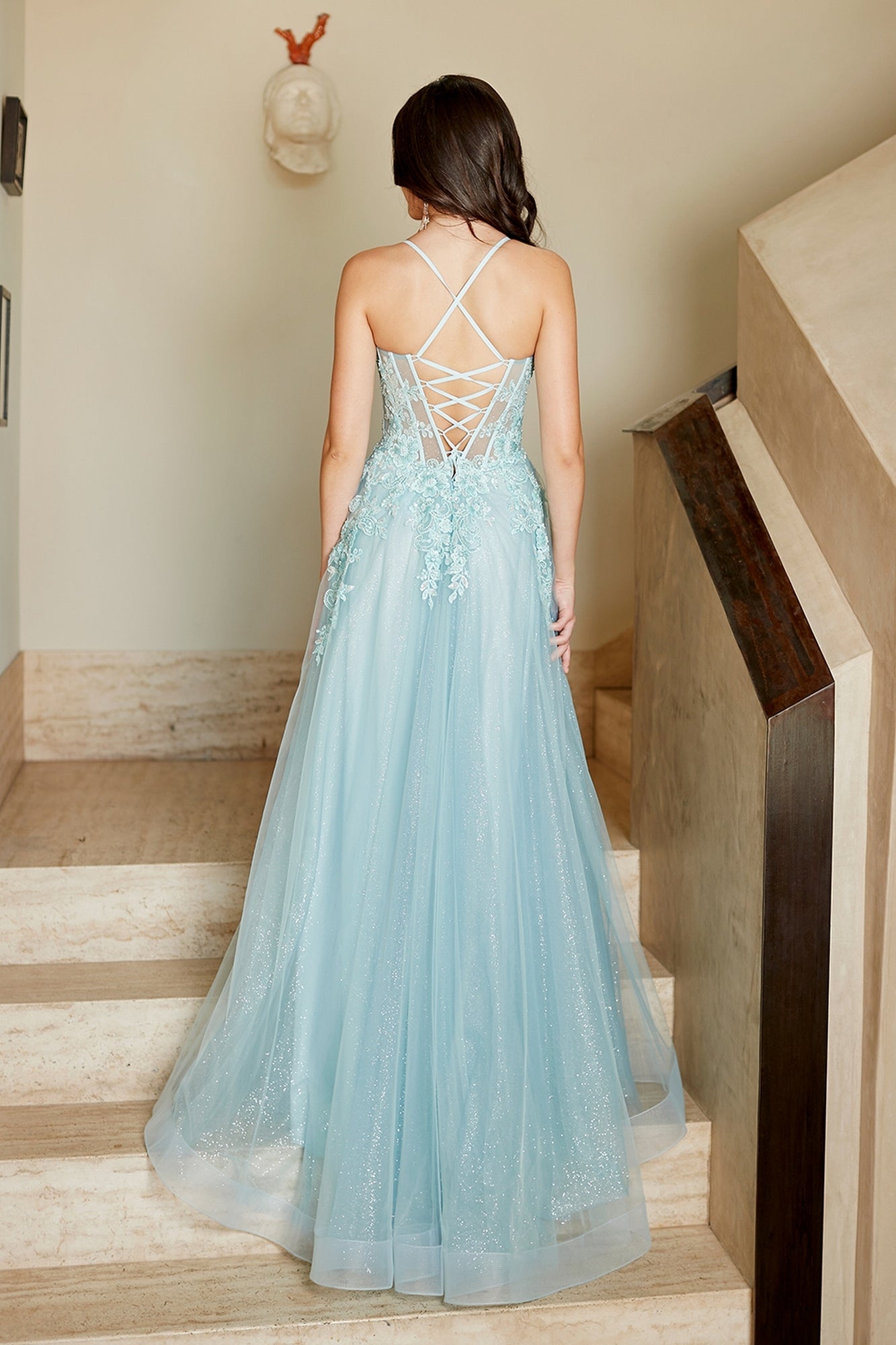 Back view of a light blue Amelia Couture 4074 floor-length gown with a lace-up corset back and floral lace bodice details, standing on a staircase.